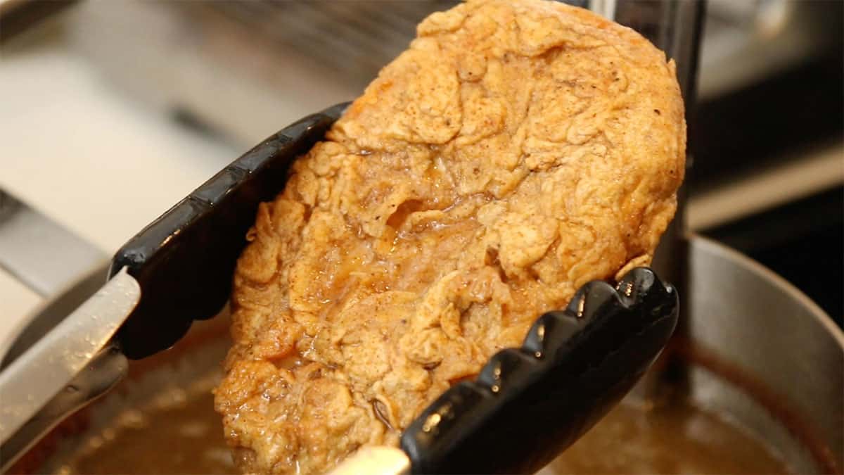a pair of tongs holding a vegan fried chicken patty over a pot of frying oil
