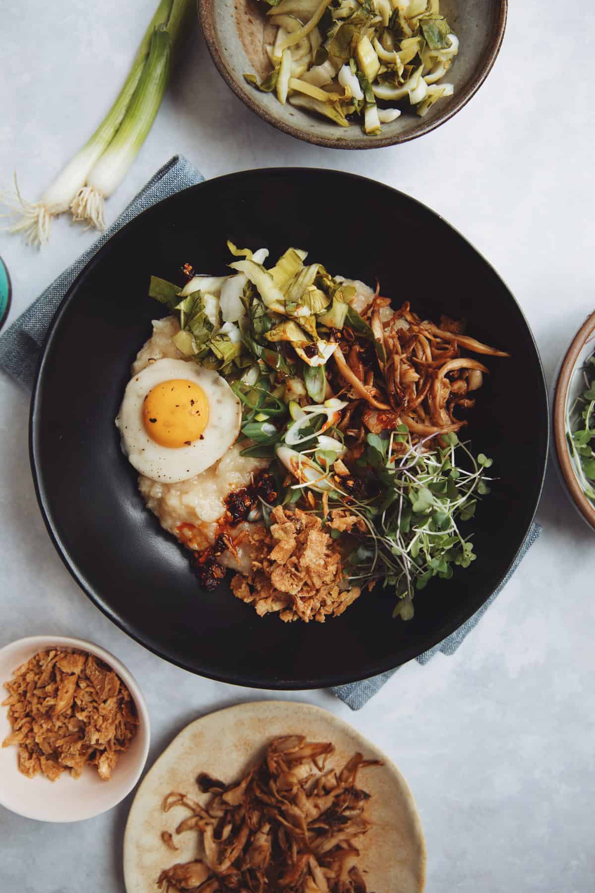 vegan breakfast congee bowl topped with shredded king oyster mushrooms, pickled bok choy, crispy fried onions, micro greens, and scallions.